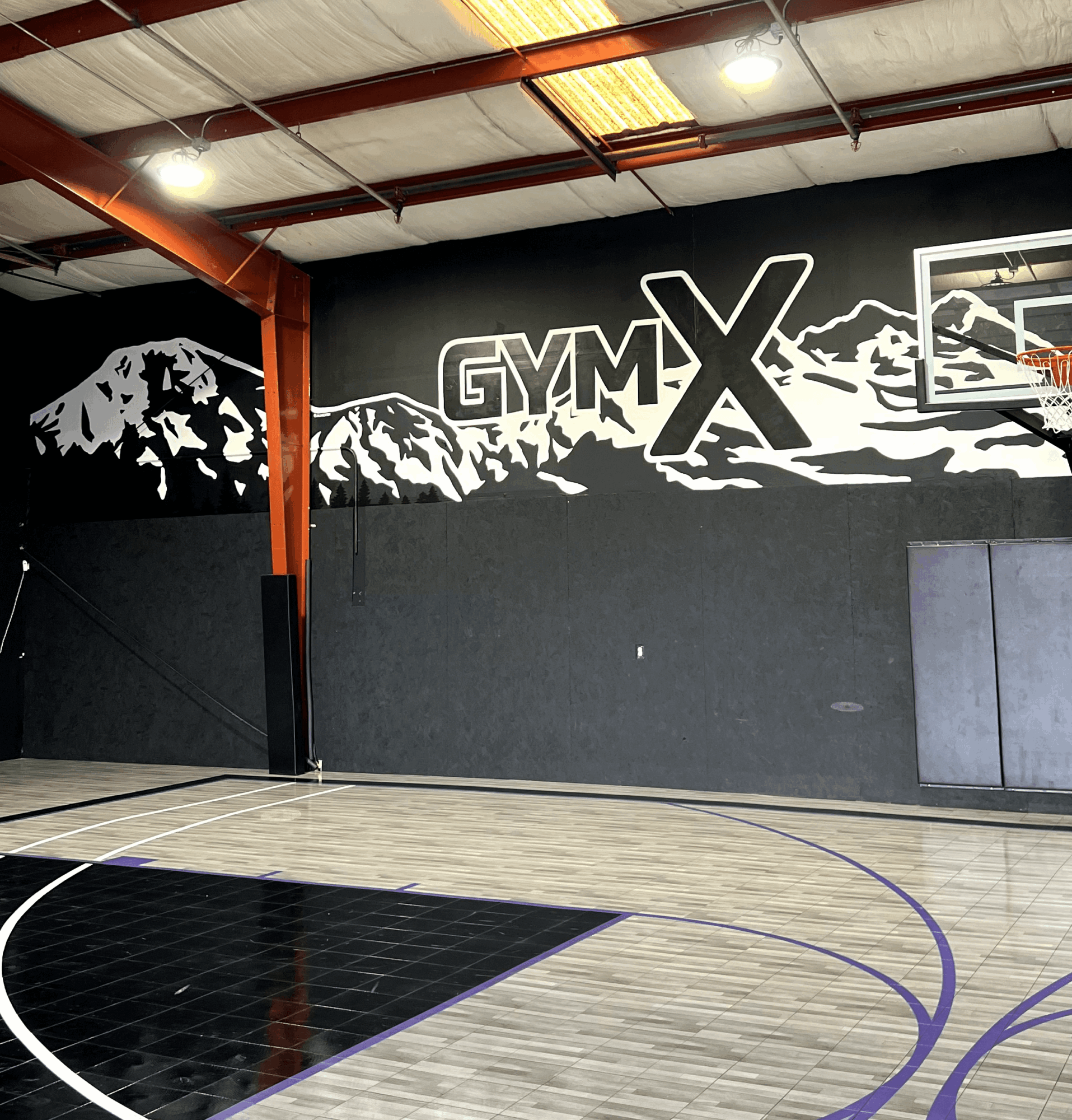 Indoor gym with a mountain mural and a basketball hoop, gymX logo on wall, wooden floor, and sports equipment.