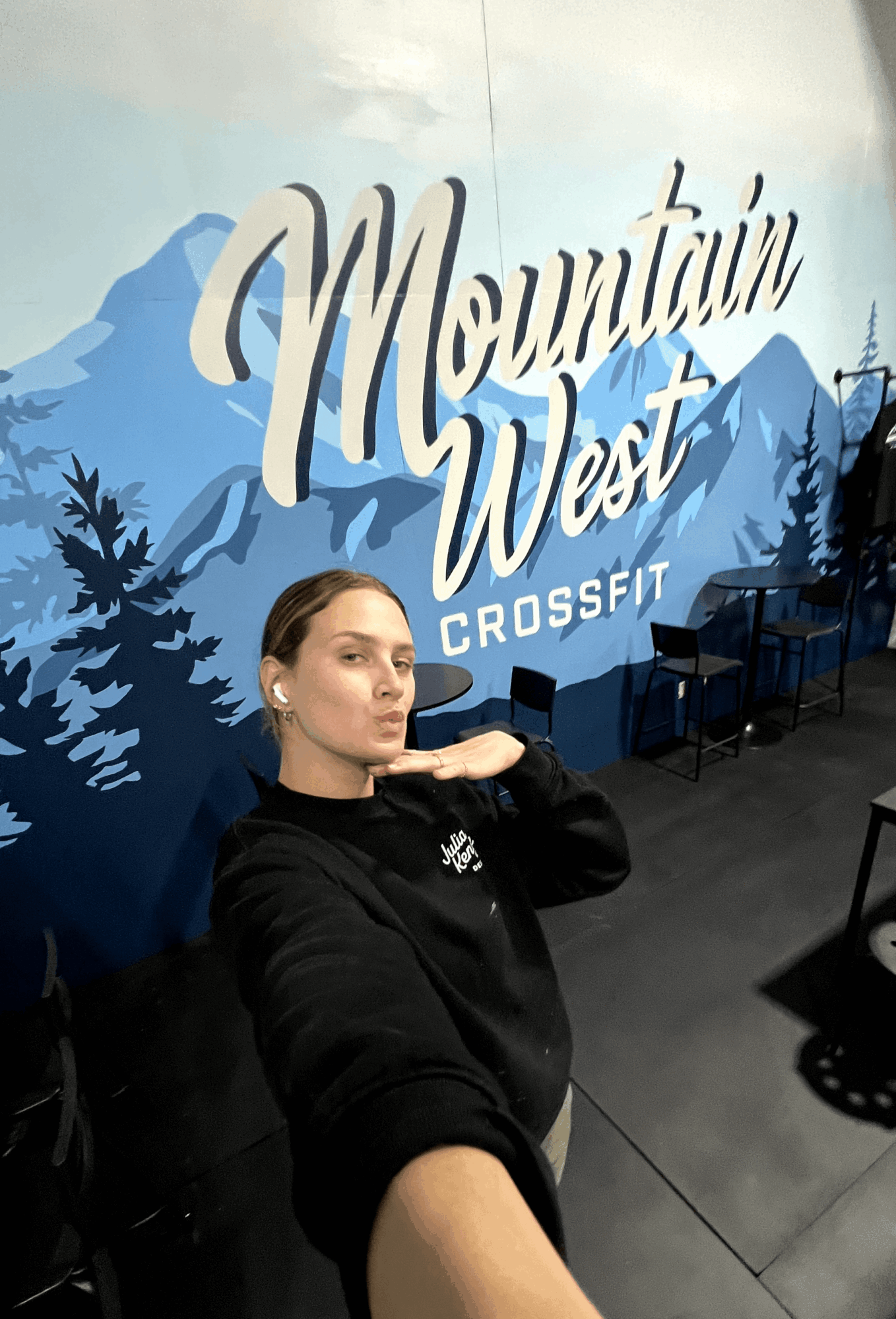 Woman taking a selfie in front of a mountain-themed wall with the words 'Mountain West CrossFit'.