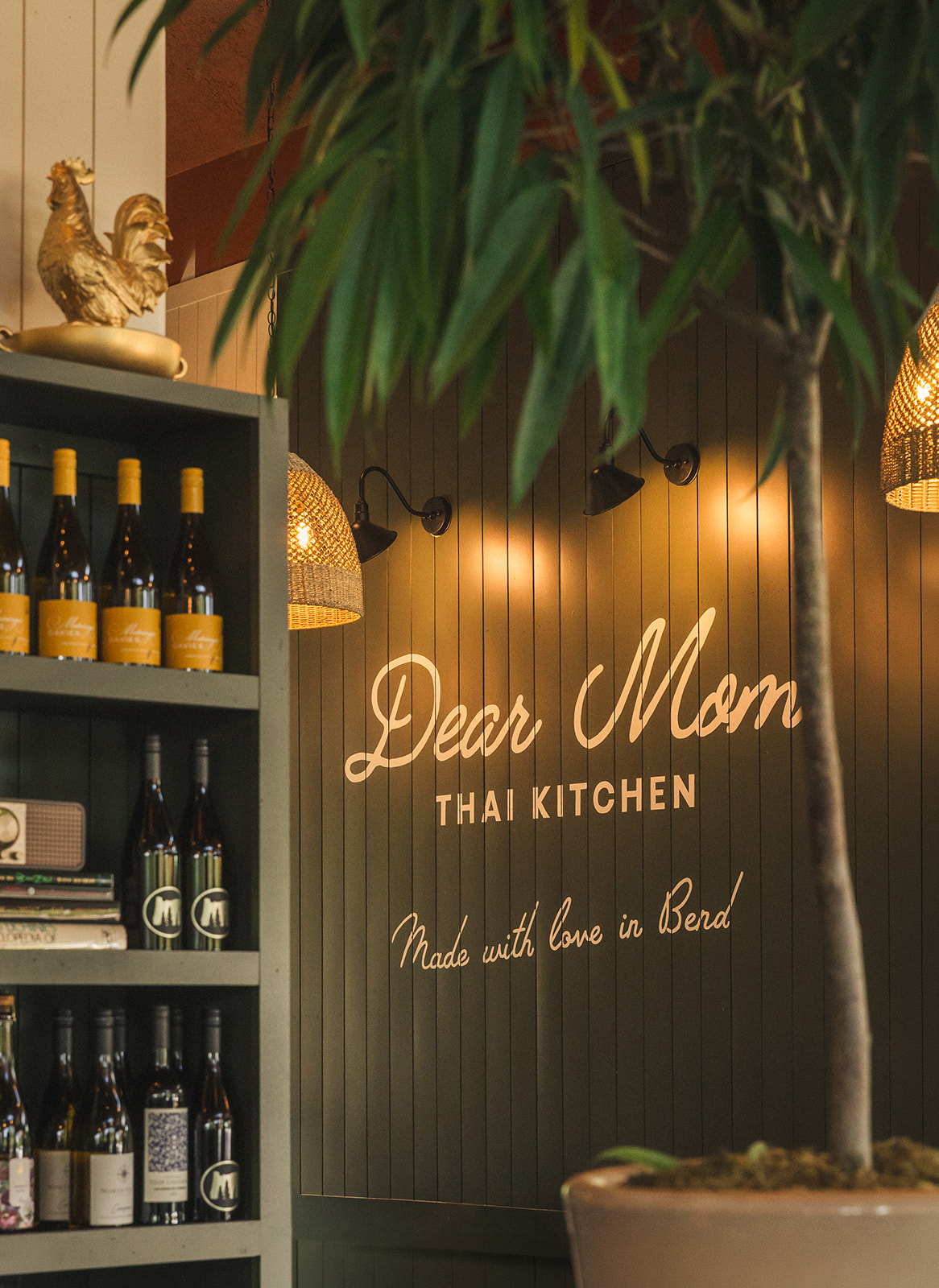 Neon sign reading 'Dear Mom Thai Kitchen' with plants and shelves of wine bottles in the background.