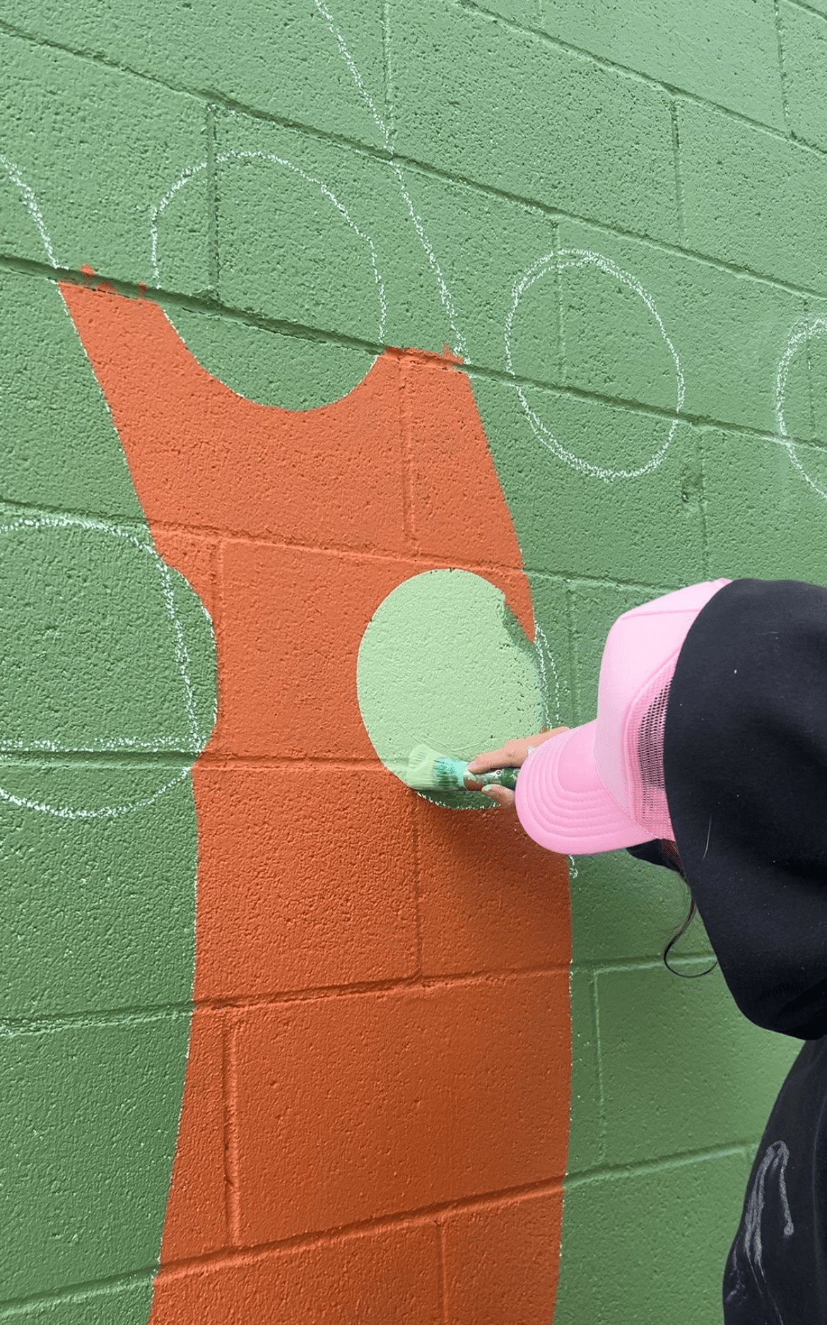 Person using spray paint on a green brick wall with orange and light green shapes, wearing a pink cap and black jacket.