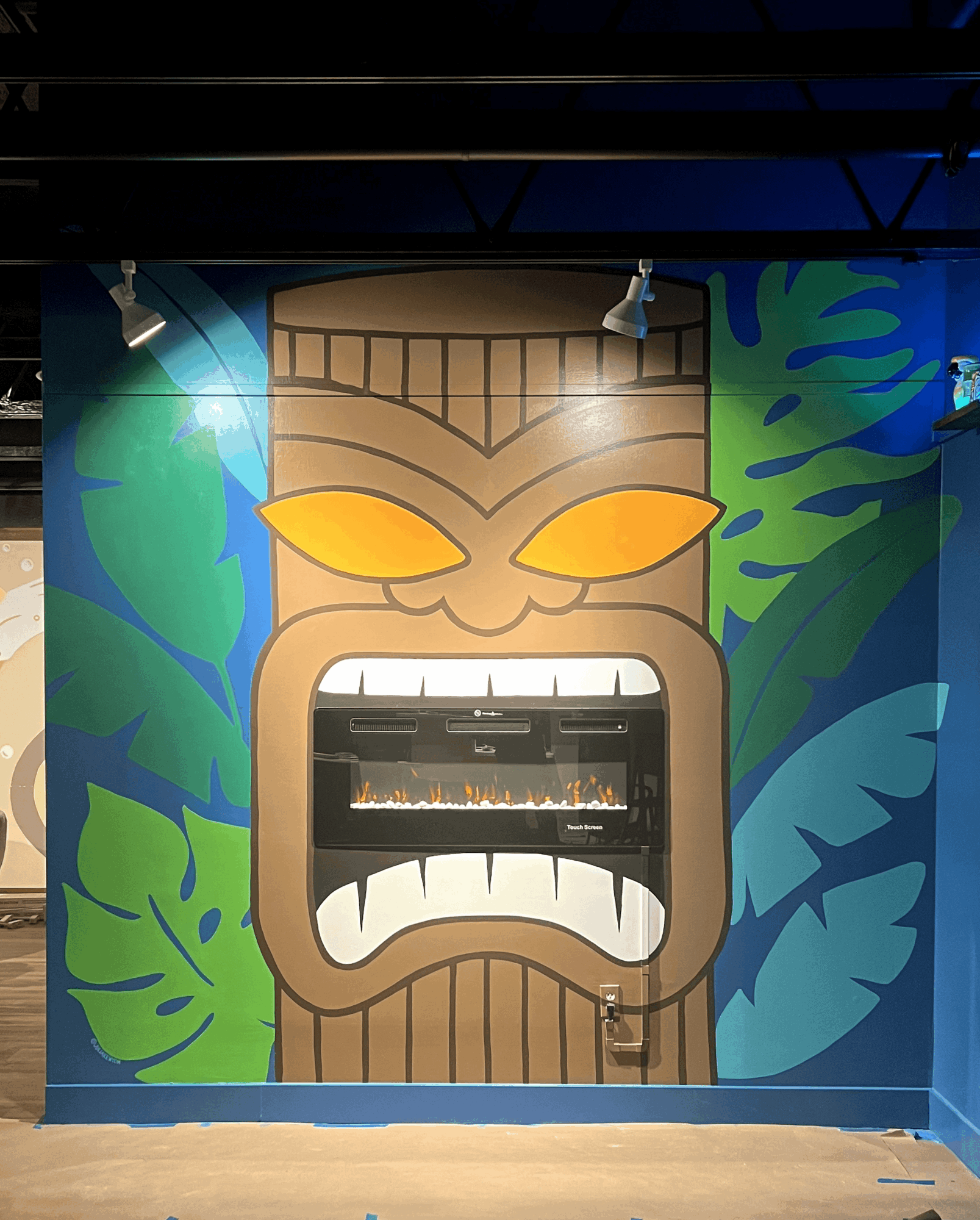 Tiki face with glowing eyes and open mouth, surrounded by green and blue foliage, on a colorful wall.