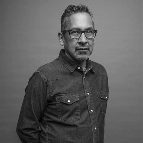 Man with glasses and short hair wearing a button-up shirt, facing slightly to the right, against a plain background.