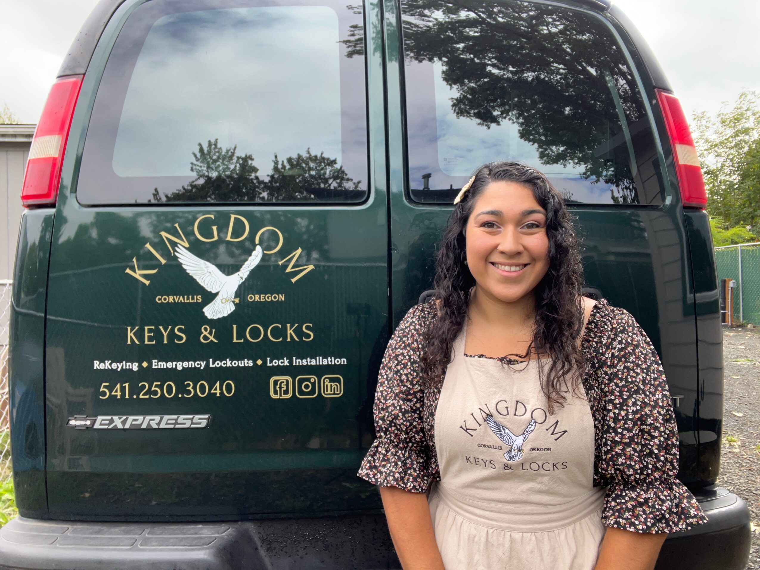 Smiling woman standing in front of a green van with company logo and contact information, outdoors with trees reflected in the windows.
