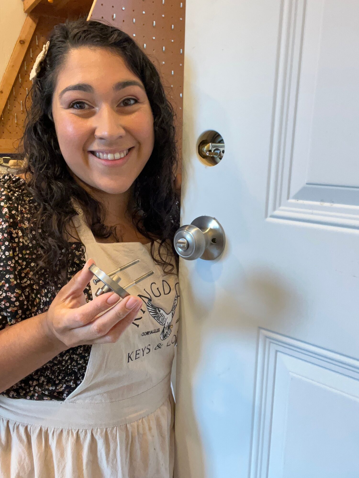 Smiling woman with dark hair taking a selfie in front of a door with a doorknob, wearing a beige top and black patterned sleeves.