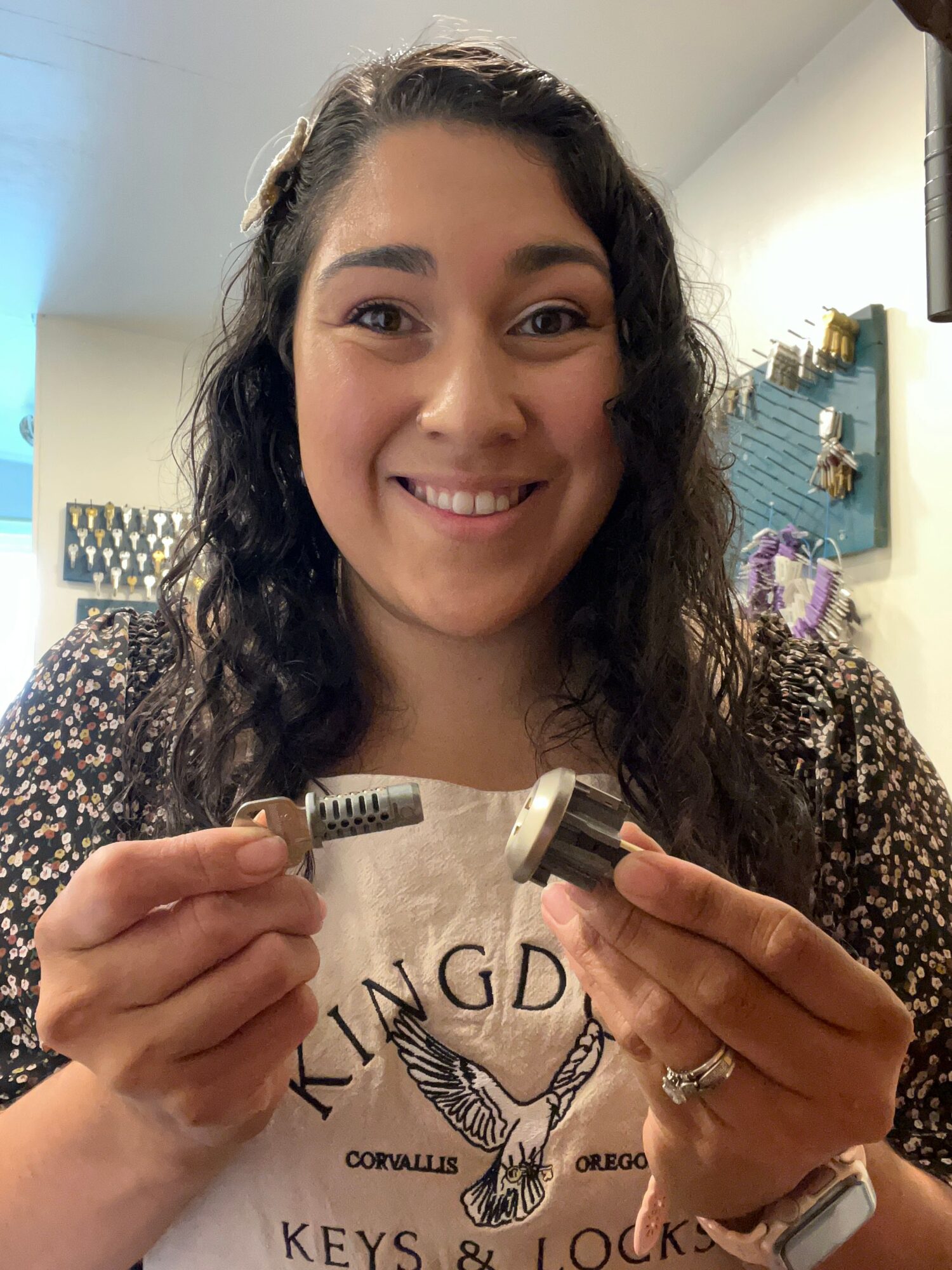 Woman with dark curly hair smiling, holding a small object, wearing a ring and a printed shirt.