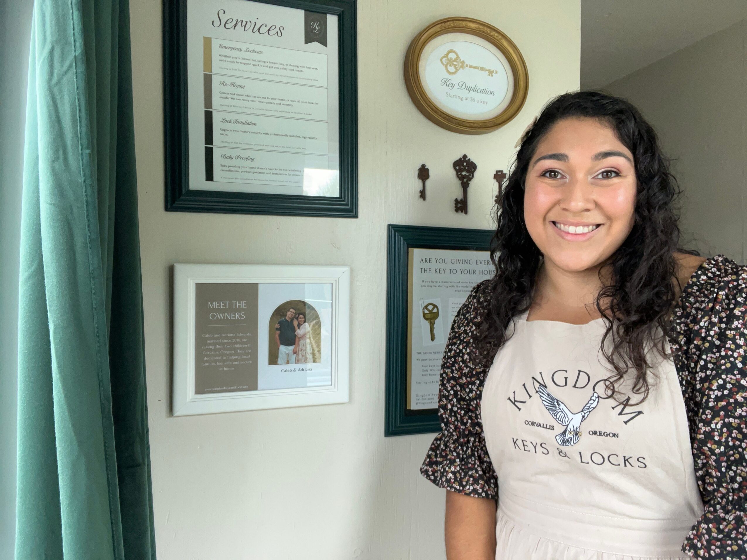 Woman with long dark hair smiling, standing indoors near framed certificates and a clock, with green curtains nearby.