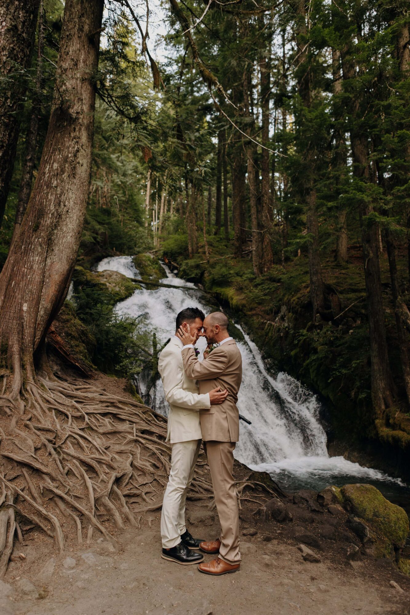 Two people embrace in front of a waterfall surrounded by trees in a forest.