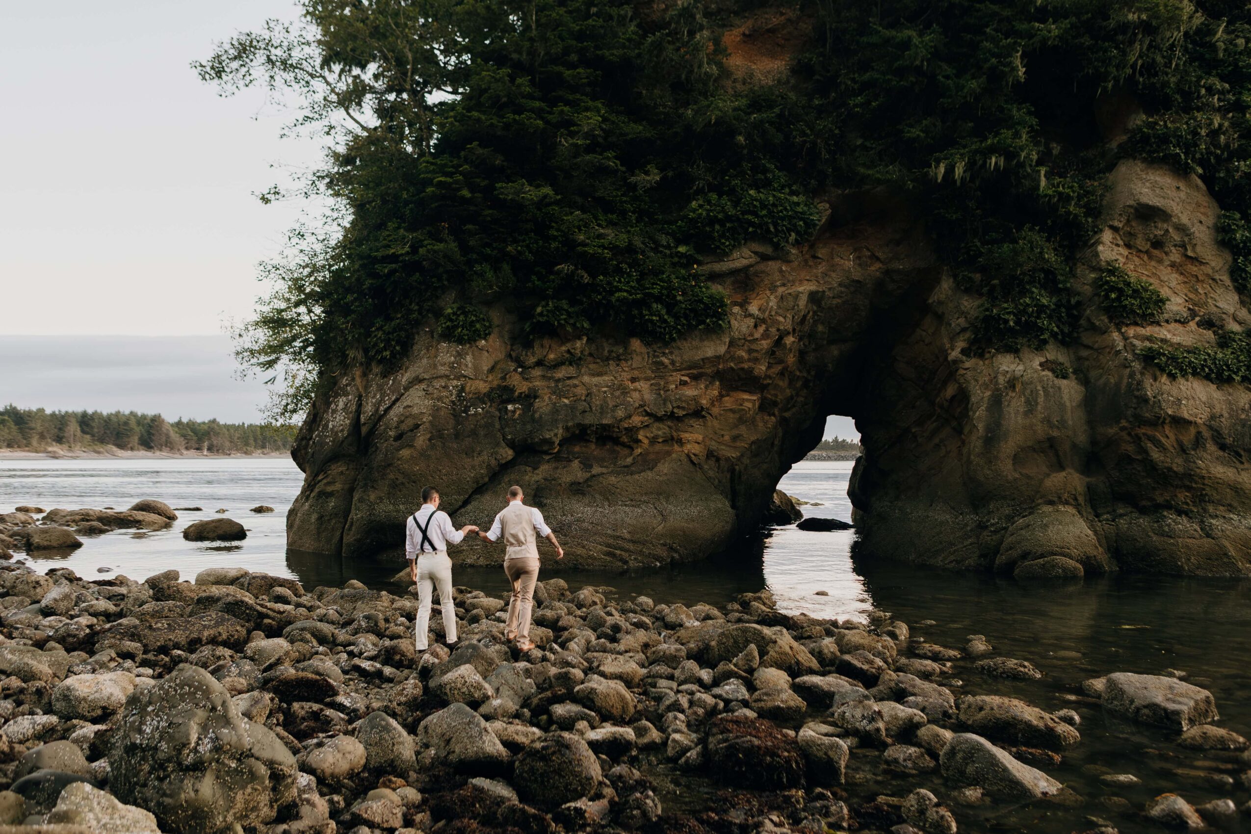 Two people stand on rocks near a large rock formation with a natural arch by the water, trees above.