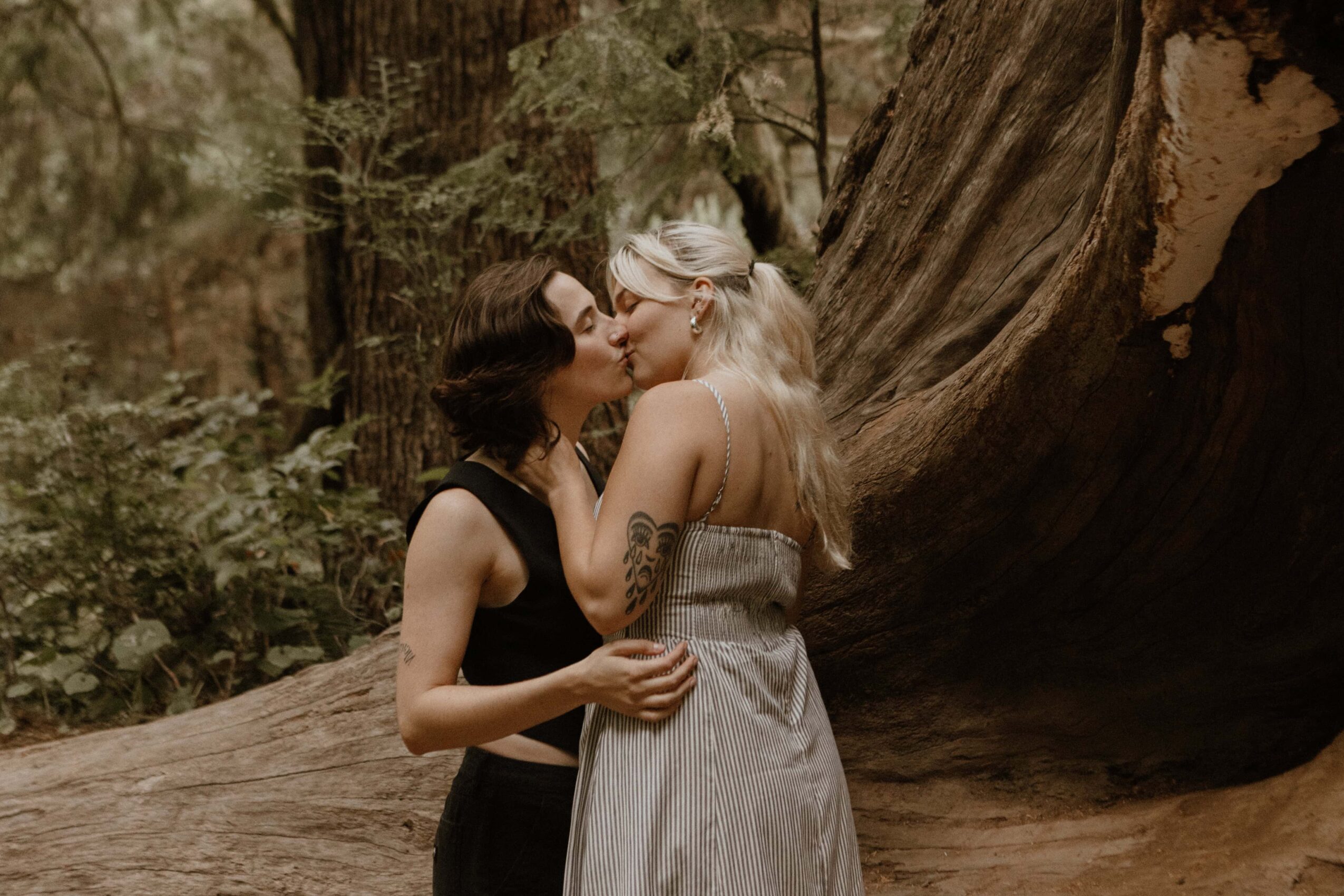Two women kissing outdoors near a large tree, one with dark hair and the other with light hair, in a forest setting.
