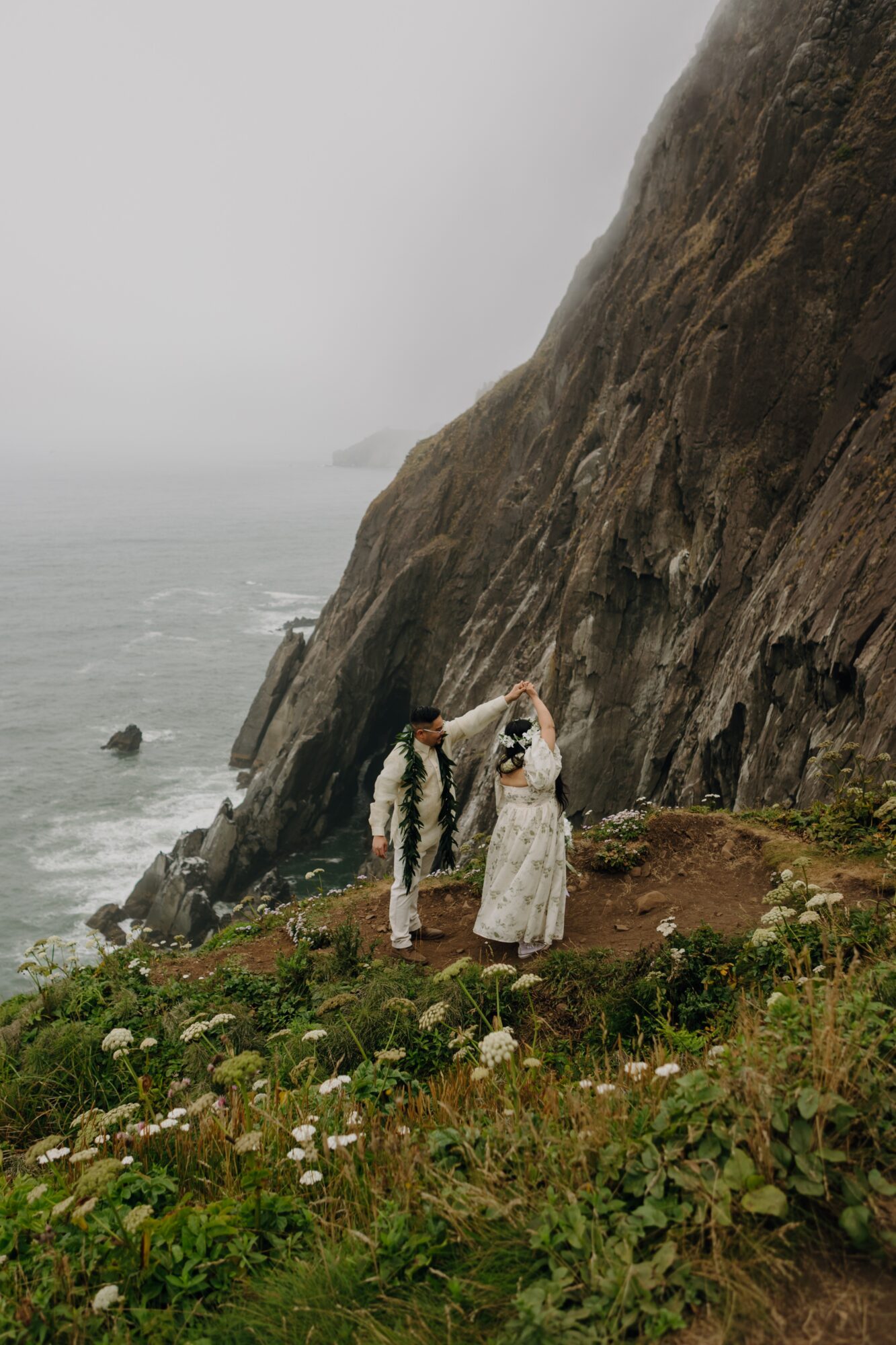 Two people stand on a grassy area near a large rocky cliff by the ocean, with foggy weather in the background.