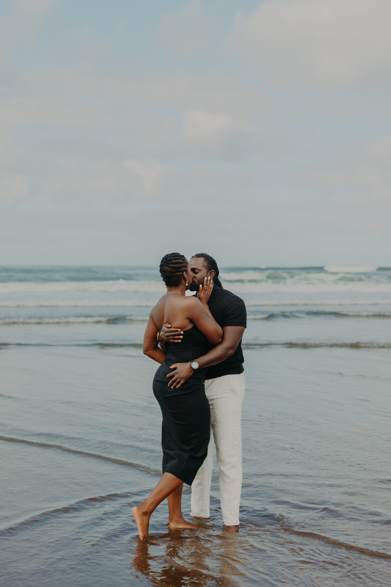 Two people stand close together in shallow ocean water, embracing and touching faces, with a cloudy sky and waves in the background.