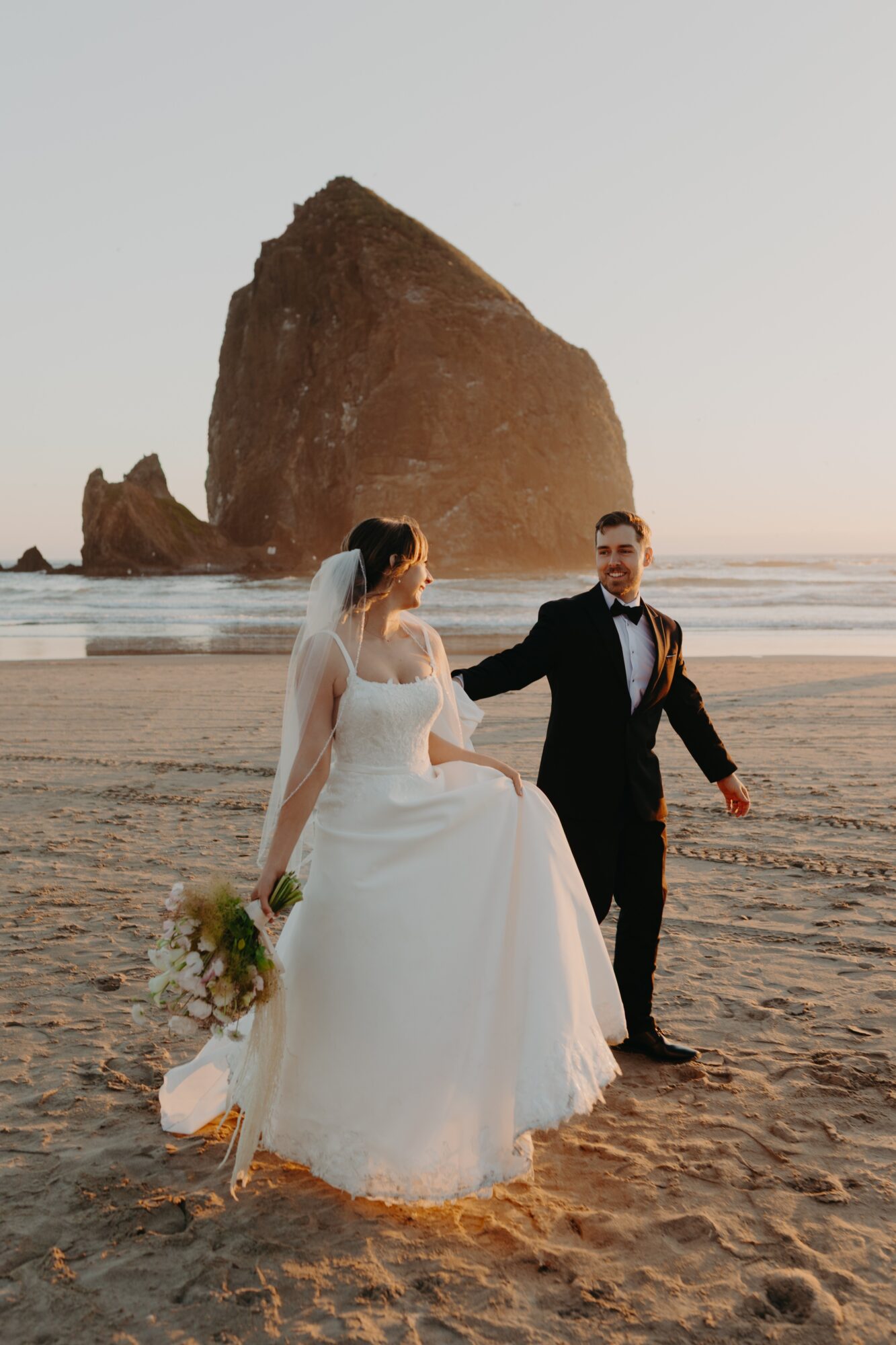 A bride and groom walk on a beach with a large rock formation in the background, holding hands and smiling.