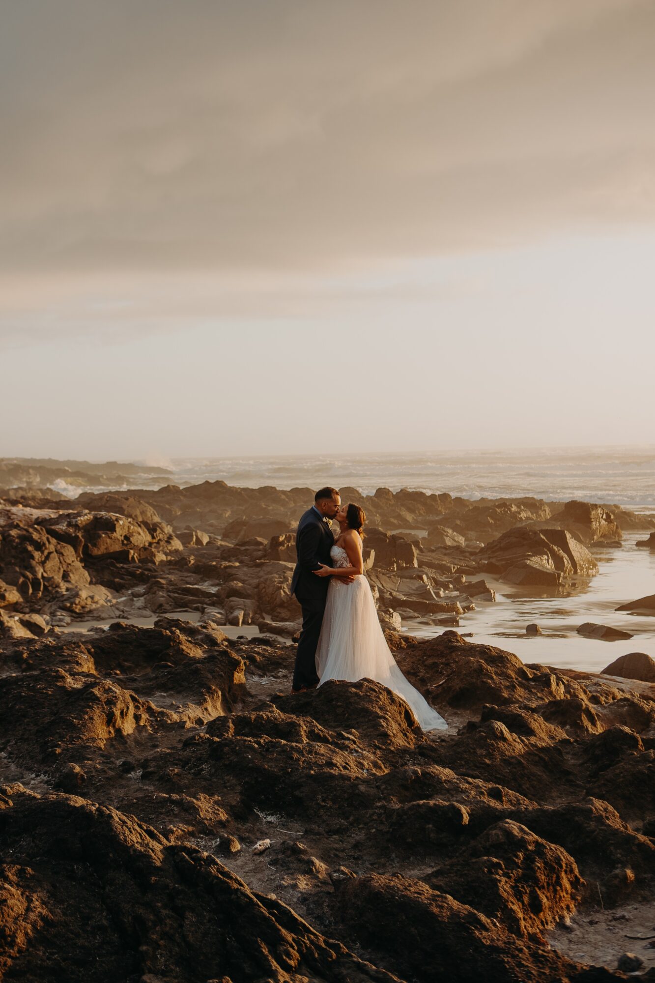 Couple in wedding attire standing on rocky beach at sunset, embracing with ocean in background.