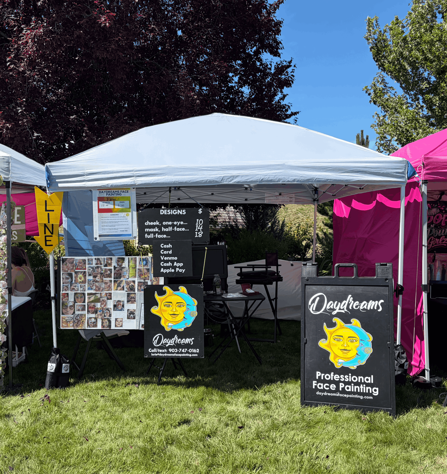 Outdoor booth with white canopy, signs for face painting, and colorful displays on grass, trees, and blue sky in background.