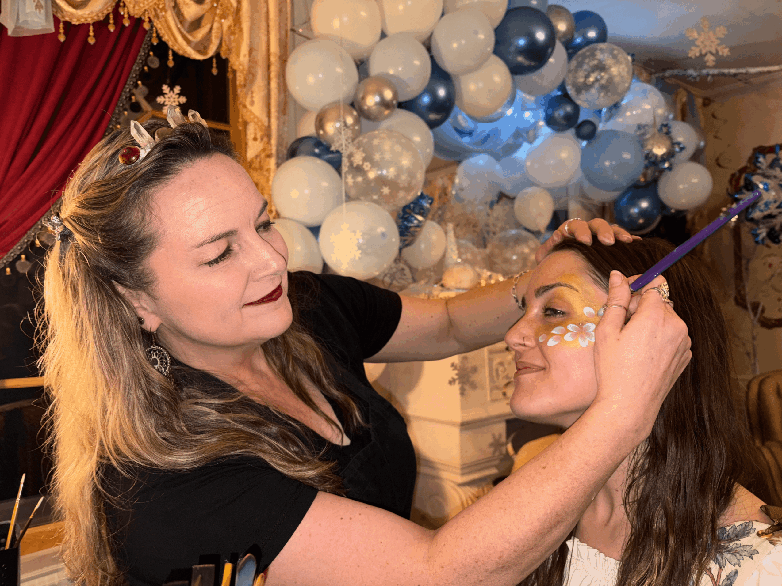 Woman applying makeup to a smiling girl in front of a decorated balloon backdrop, celebrating a festive event.