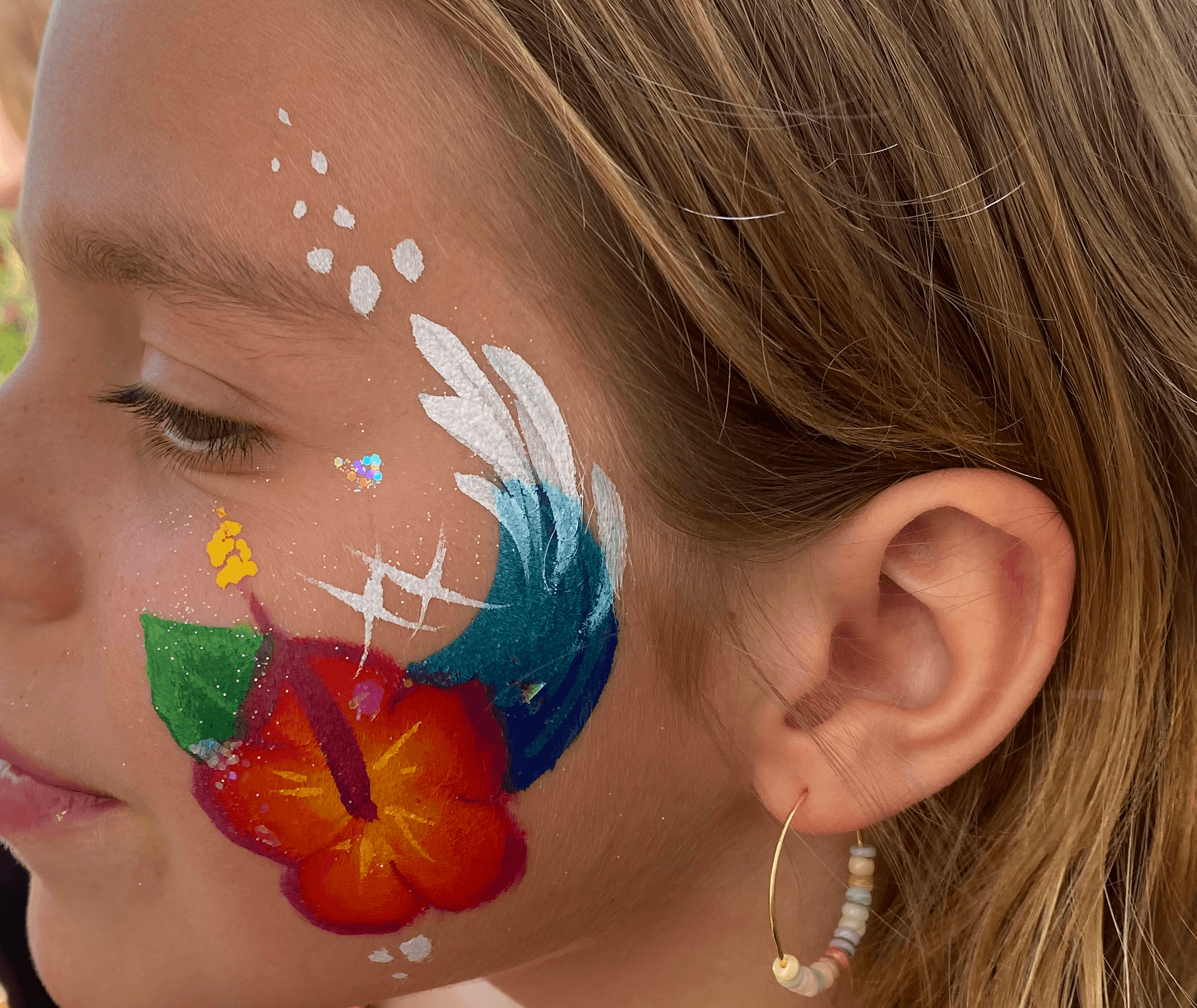 Child with face paint of a flower, leaves, and abstract designs on cheek, wearing a beaded earring, with closed eyes.