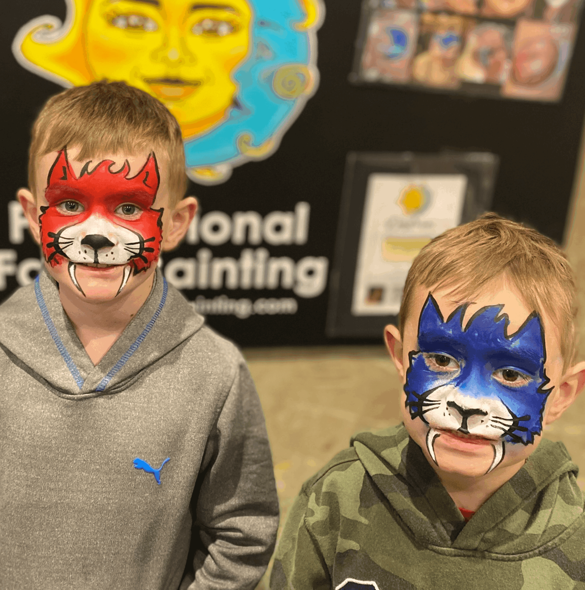 Two children with face paint of cartoon cats, one red and one blue, standing indoors with a colorful background.
