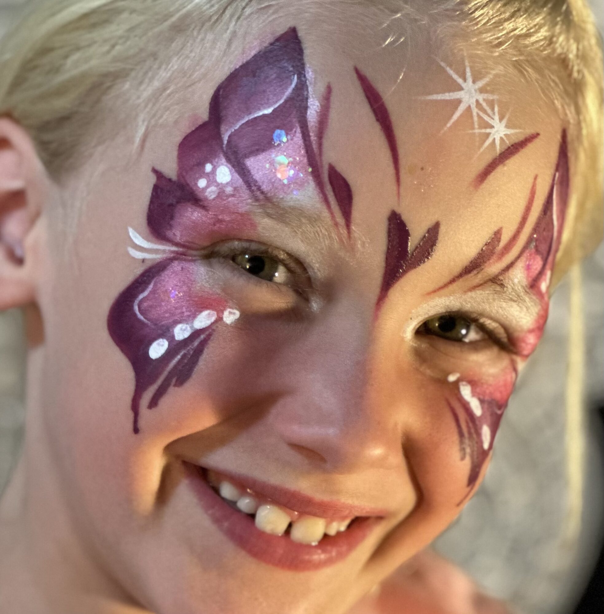 Child with colorful butterfly face paint, smiling, showing missing teeth, with star and swirl designs around eyes.