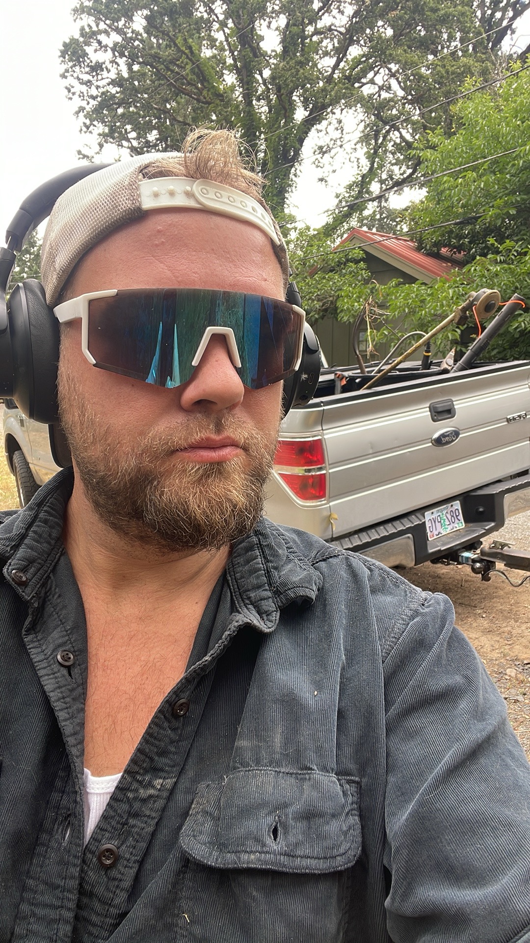 Man wearing sunglasses, headphones, and a cap outdoors with a truck and trees in background.