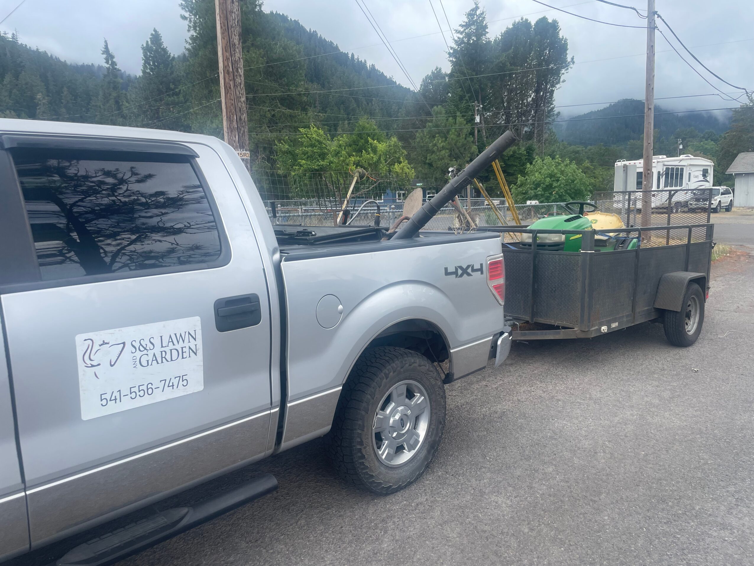 Silver pickup truck with a trailer attached, parked outdoors with trees and power lines in the background.