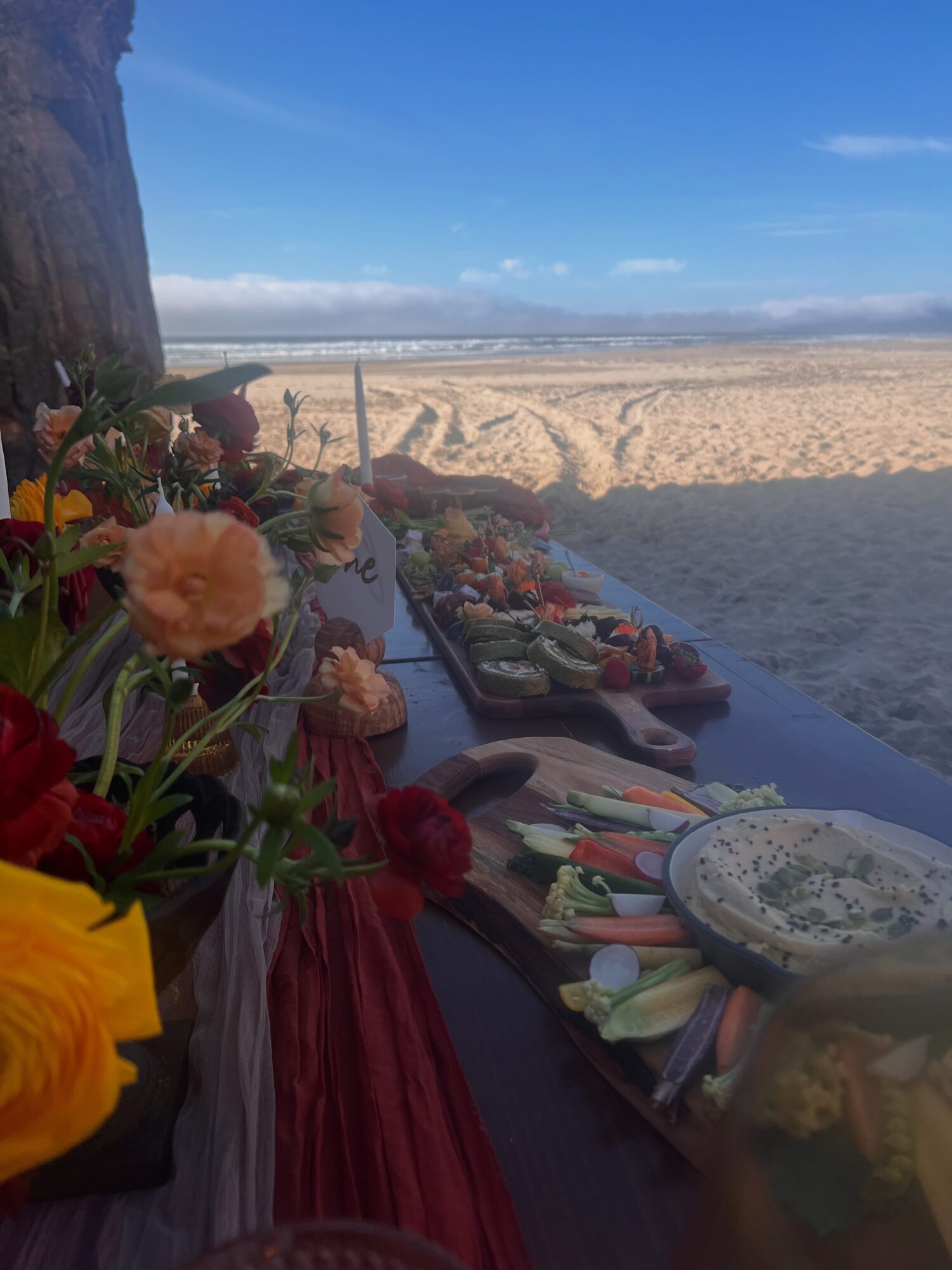 Beach scene with a table of food and flowers, sandy beach, blue sky, and ocean in the background.