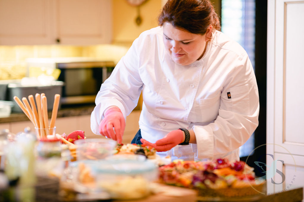 Woman in white chef's coat preparing food in a kitchen with baked goods on the counter.