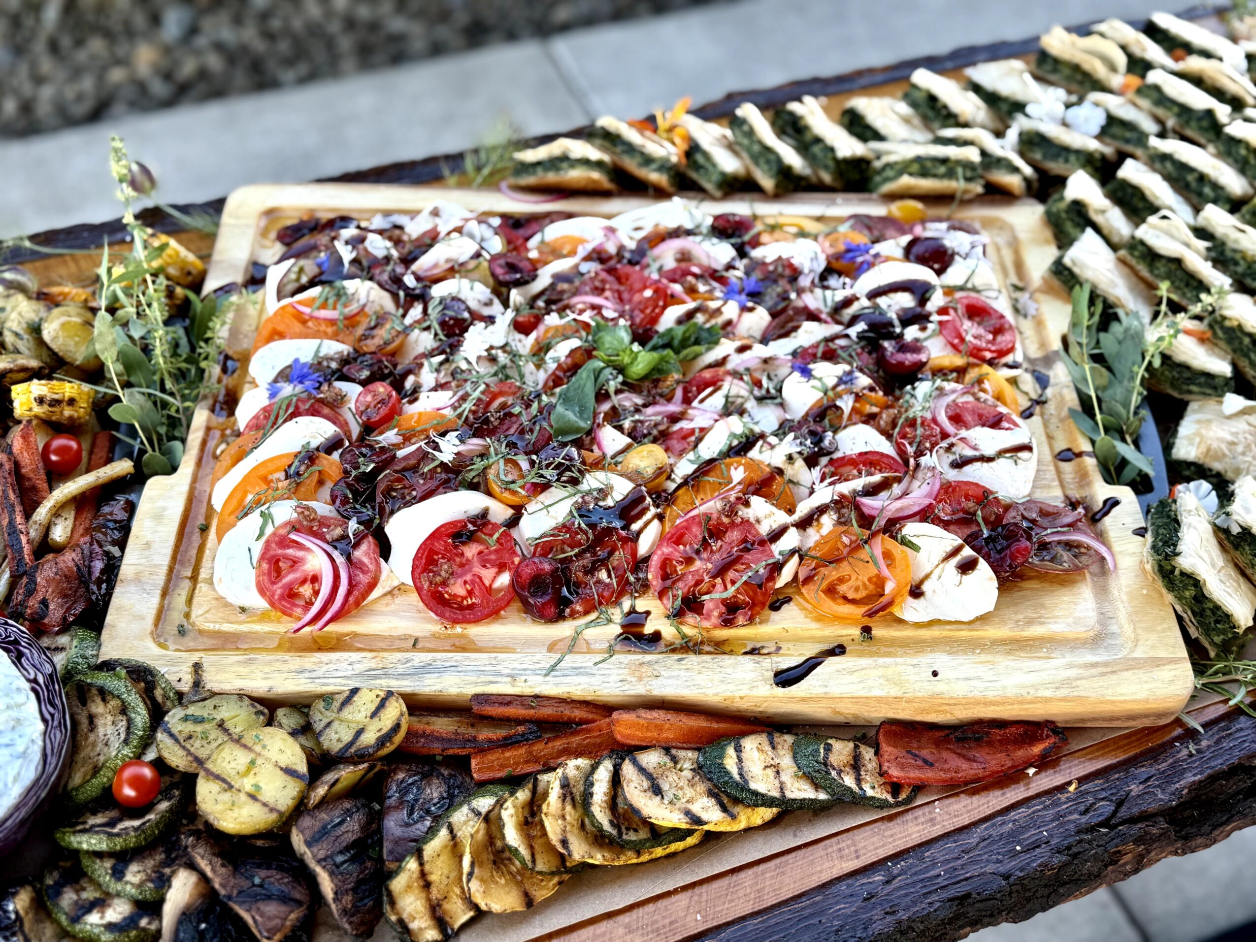 Assorted sliced appetizers on a wooden platter, garnished with herbs, set on a rustic table outdoors.