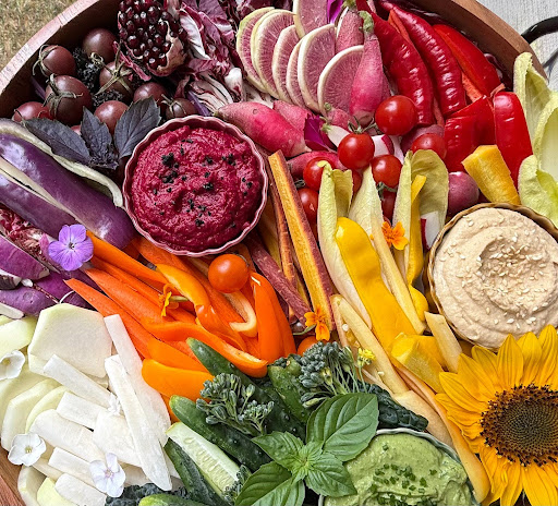 Colorful vegetable and fruit platter with cherry tomatoes, radishes, carrots, cucumbers, hummus, and sunflower, arranged in a circular pattern.