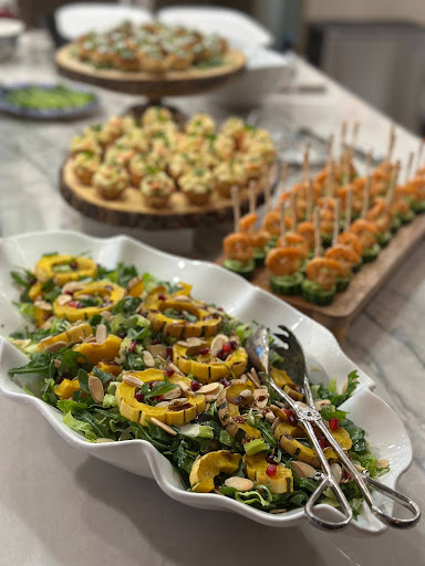 Assorted appetizers on a table, including skewers, salads, and small bites, with a blurred background of a kitchen or dining area.