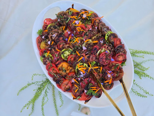 Colorful skewers with grilled meat, vegetables, and garnishes on a white oval plate, with green foliage nearby.