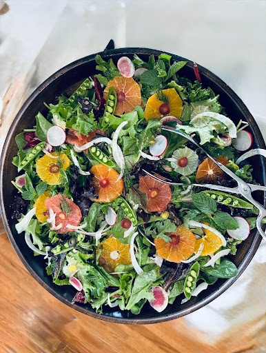 Colorful mixed salad with greens, orange slices, radishes, and other vegetables in a black bowl, viewed from above.