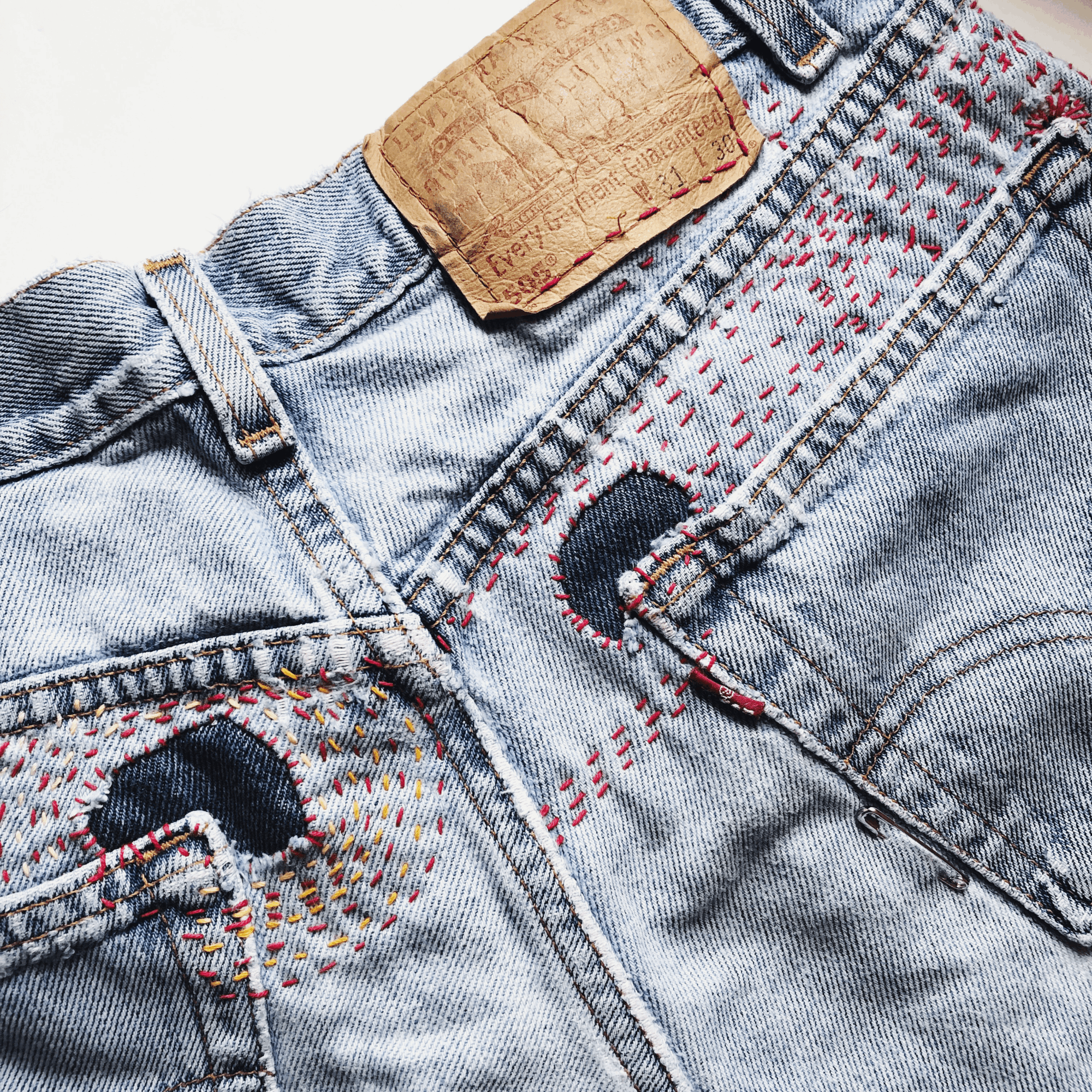 Close-up of denim jeans with a leather patch, colorful stitching, and metal rivets, showing front pockets and waistband.