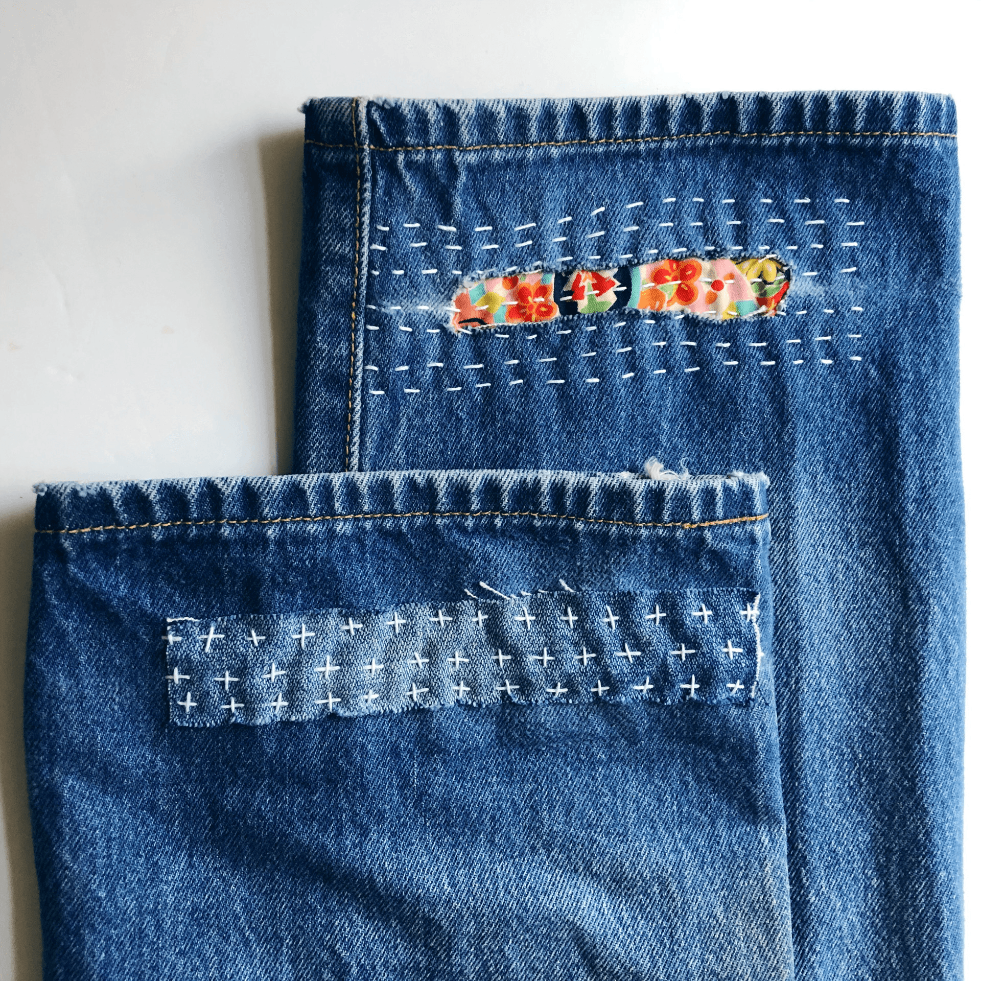 Two denim jeans pockets with decorative stitching and embroidery, one with floral embroidery, on a white background.