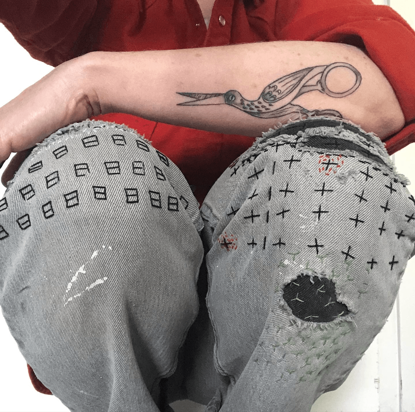 Person with a tattoo of scissors on their forearm, wearing ripped jeans with holes and patches, sitting with knees up.
