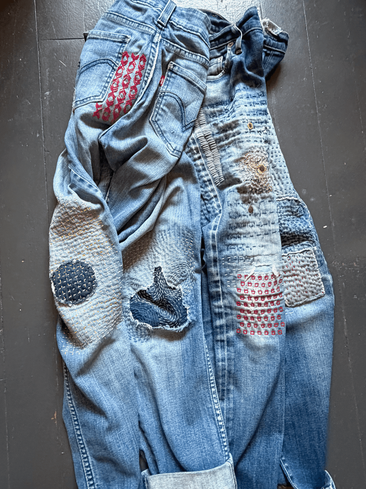 Pair of patched, distressed jeans with various colorful patches and designs, laid flat on a dark surface.