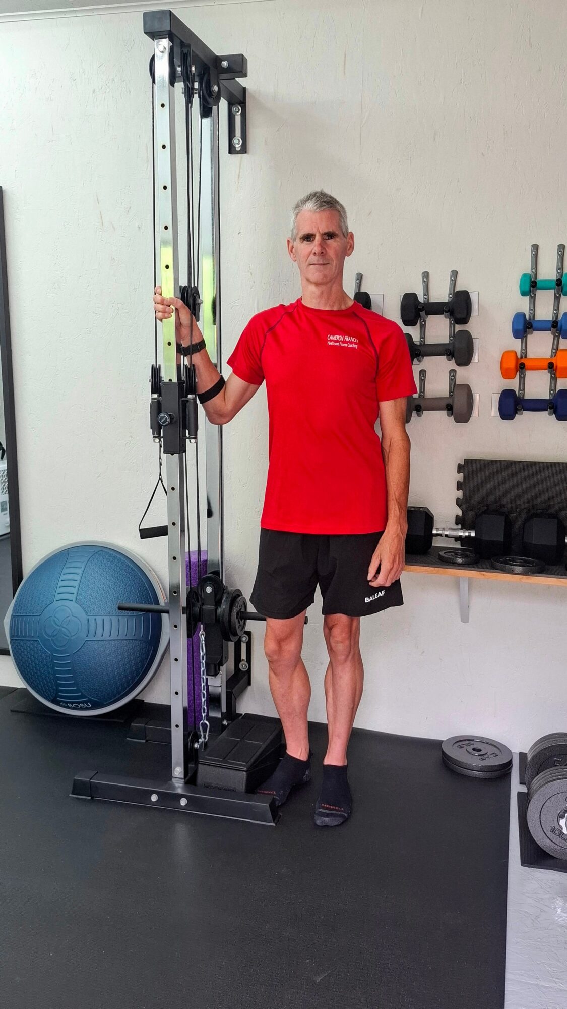 Man in red shirt and black shorts standing next to a tall exercise machine in a gym, holding a yellow resistance band.