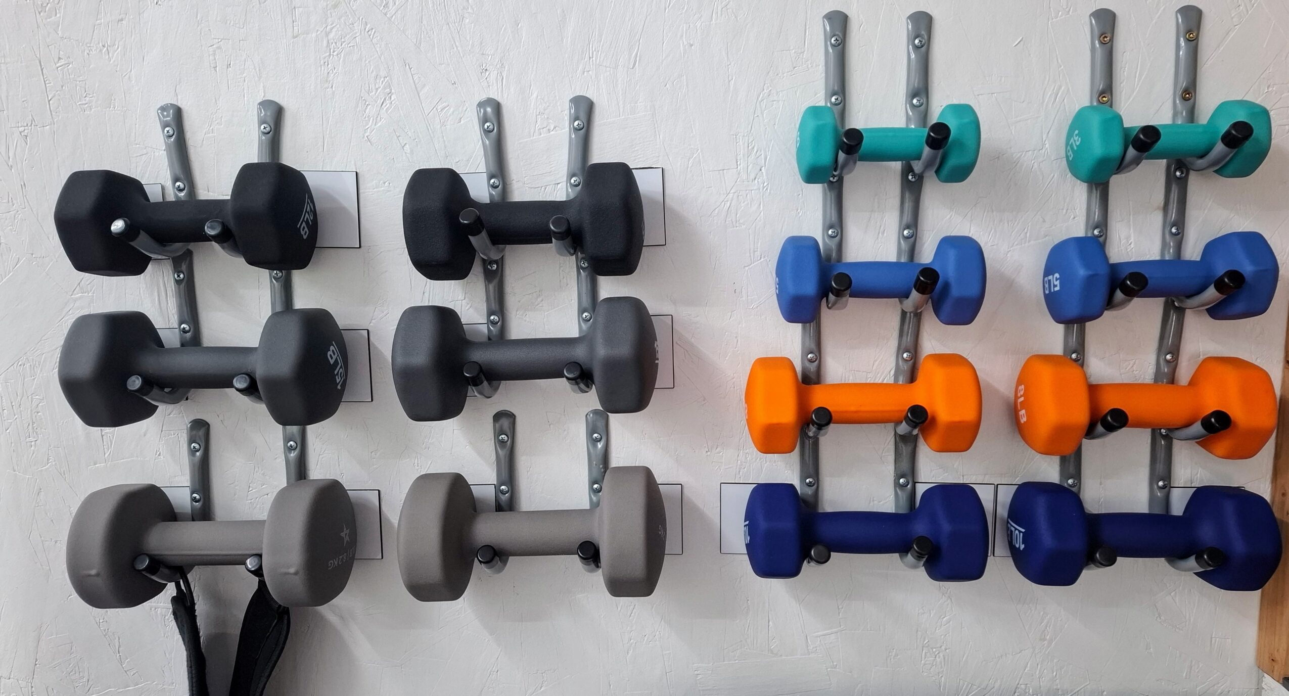 Dumbbells arranged on a wall rack in three columns with different colors, including black, gray, blue, orange, and teal.