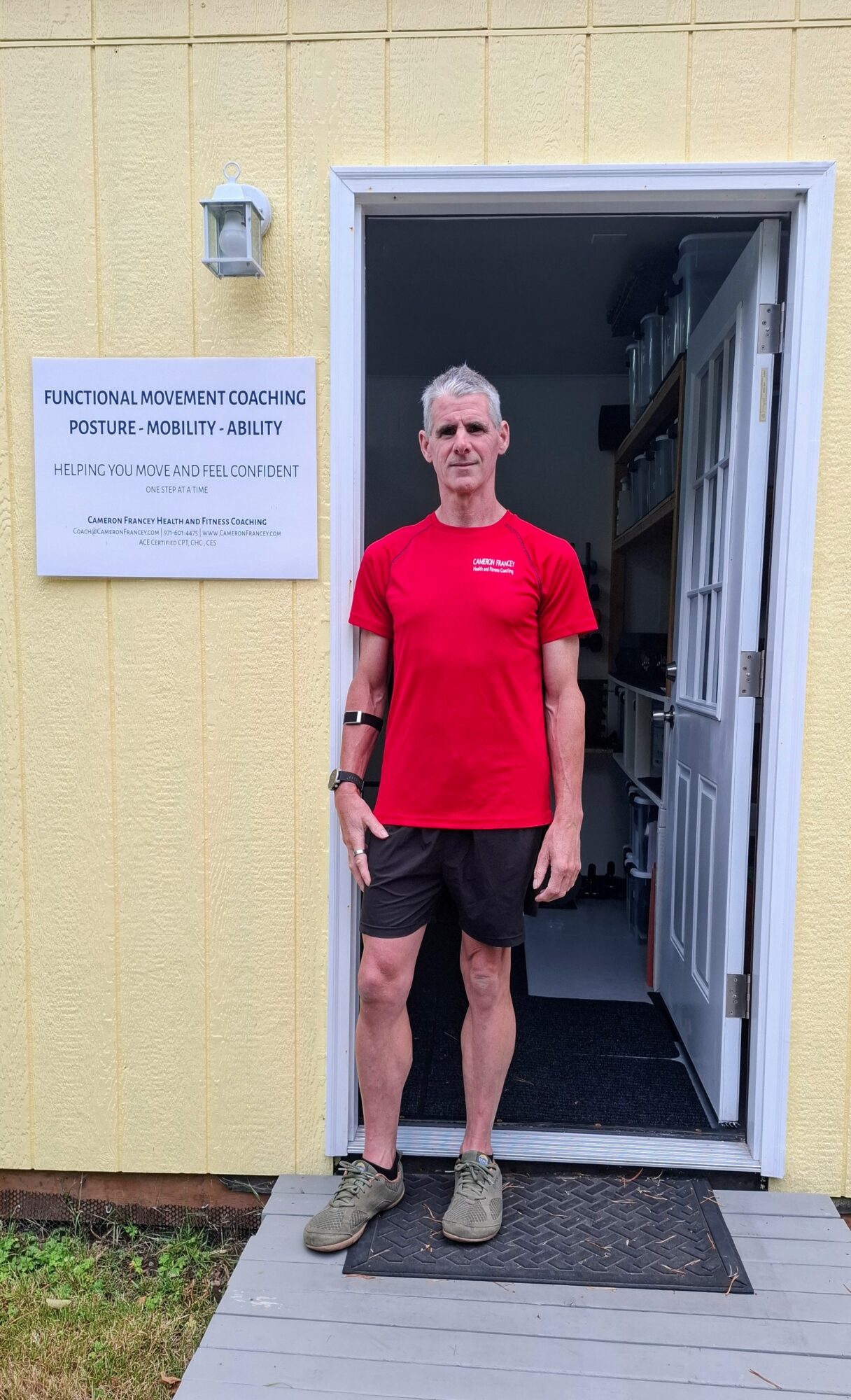 Man in red shirt and shorts standing in doorway outside a yellow building with a sign, on a gray mat.