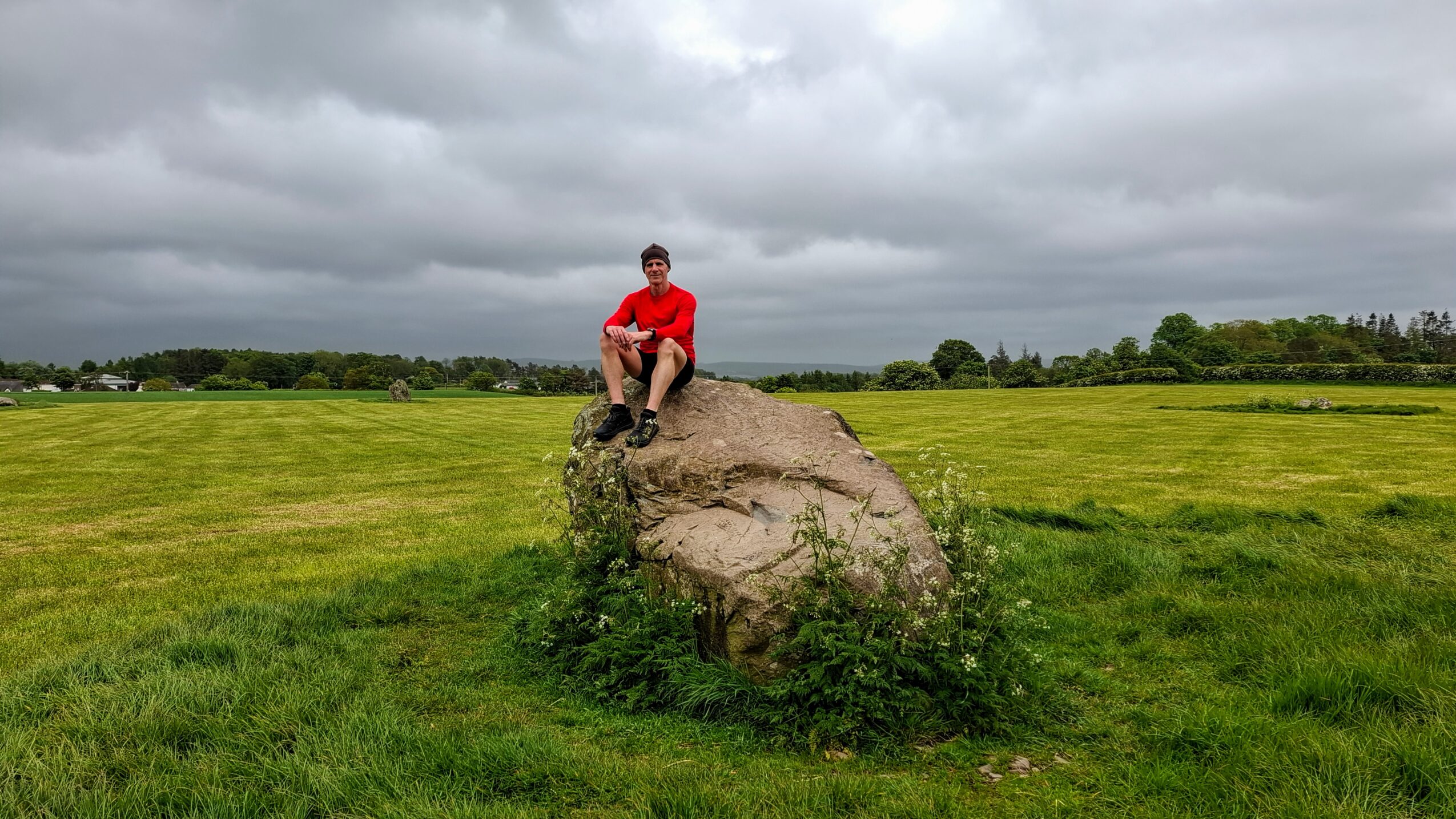 Person sitting on a large rock in a grassy field under cloudy sky.