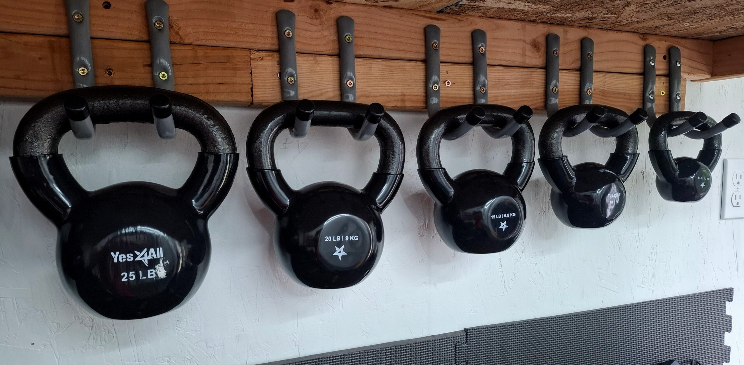 Five black kettlebells hanging on hooks on a wooden wall, with labels and a white background below.