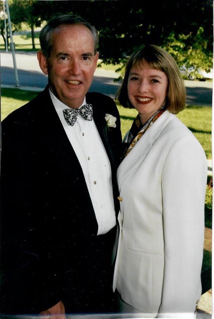 Two people, a man and a woman, standing outdoors, smiling, with trees and grass in the background.