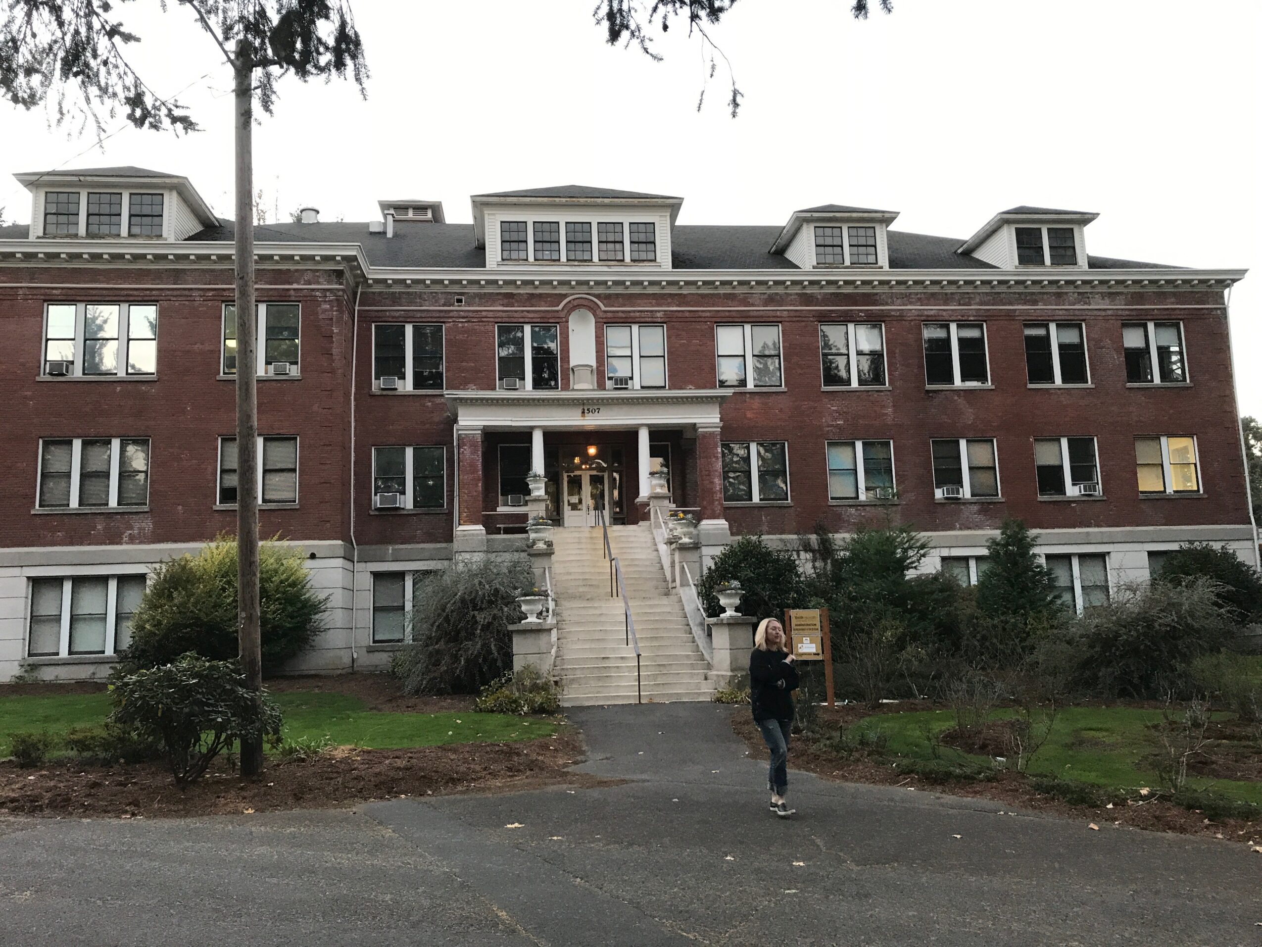 Three-story brick building with stairs leading to entrance, person walking on sidewalk, trees and bushes in front.