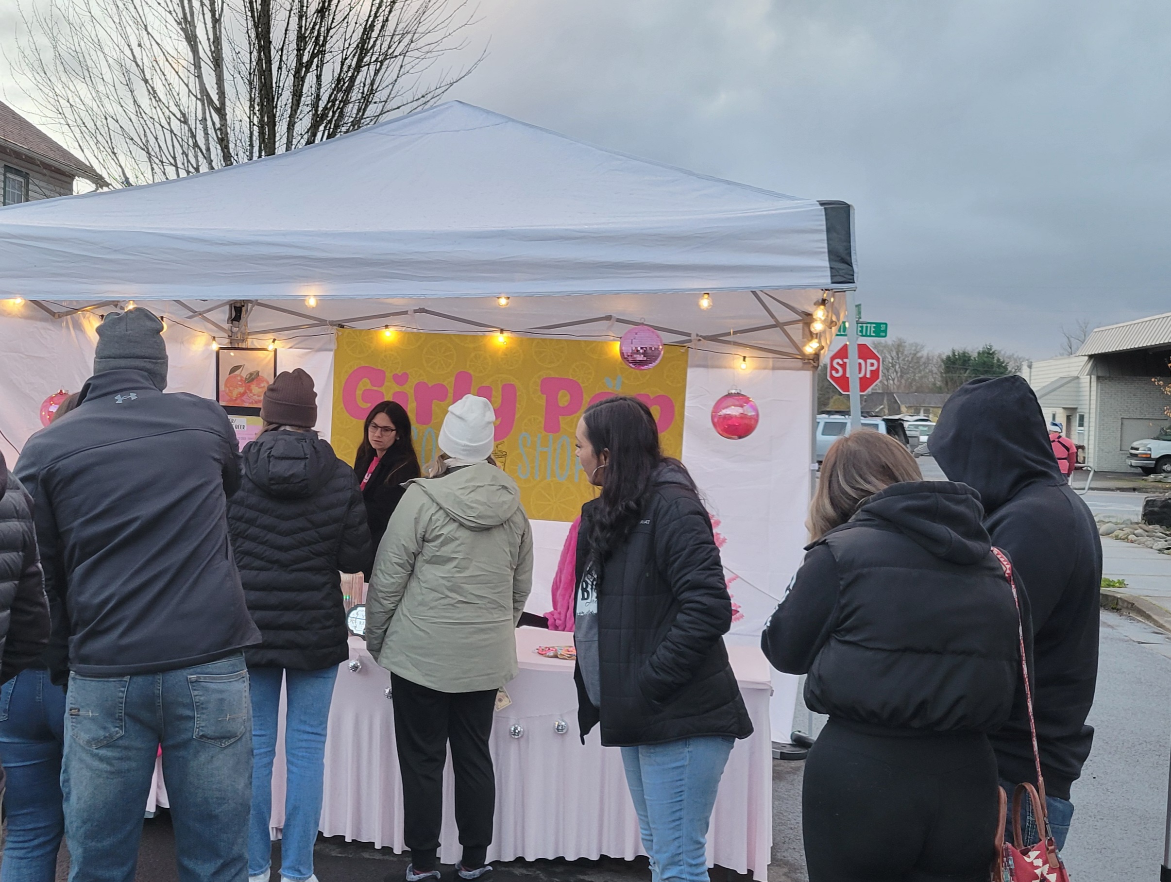 People gather at a booth under a white canopy with a pink sign, some wearing jackets and hoodies, on a cloudy day.