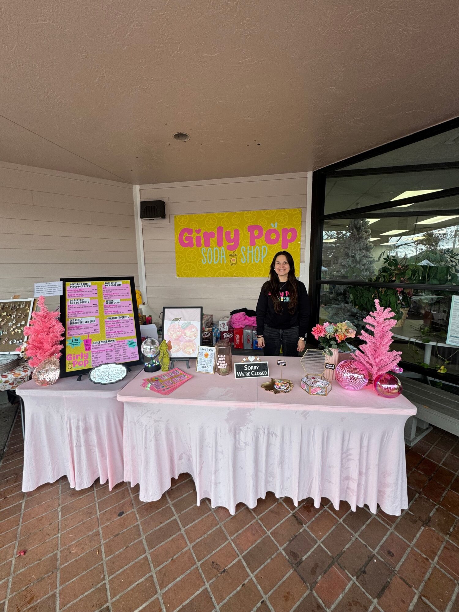 A woman stands behind a table with pink decorations and a Girl Pop sign, inside a room with brick wall and window.