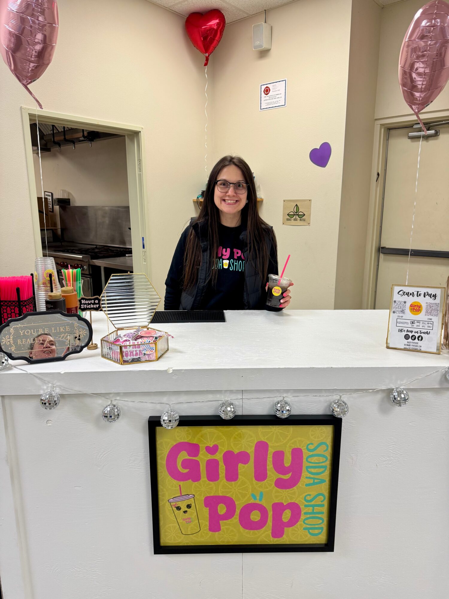 Young woman with glasses and dark hair behind a counter, holding a drink, decorated with heart-shaped balloons and purple hearts on the wall.