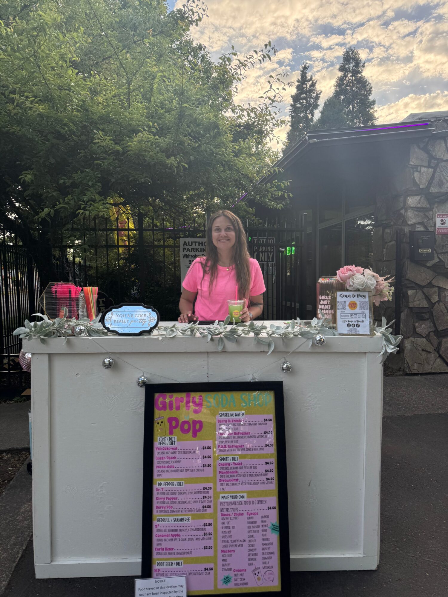 A woman in a pink shirt stands behind a white display with a colorful menu, outdoors with trees and a fence.
