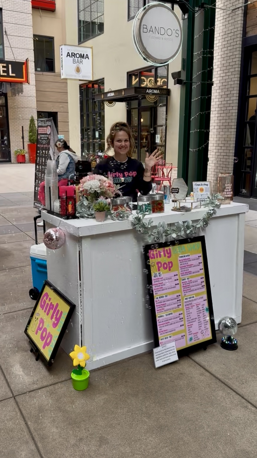 Person standing behind a white cart with flowers and pink bottles, on a city sidewalk, smiling.