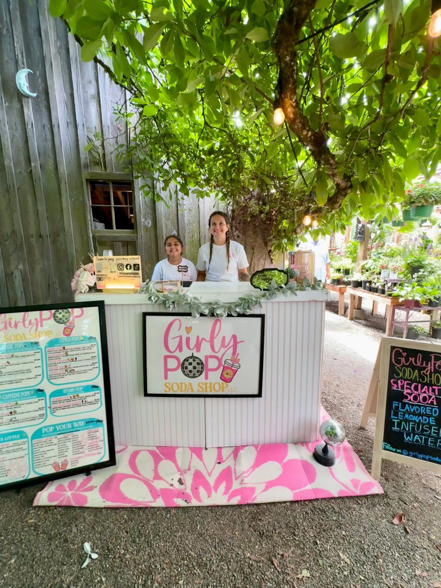 Two girls stand behind a white counter with a Girl Scout logo, under a large leafy tree, with a pink floral mat in front.