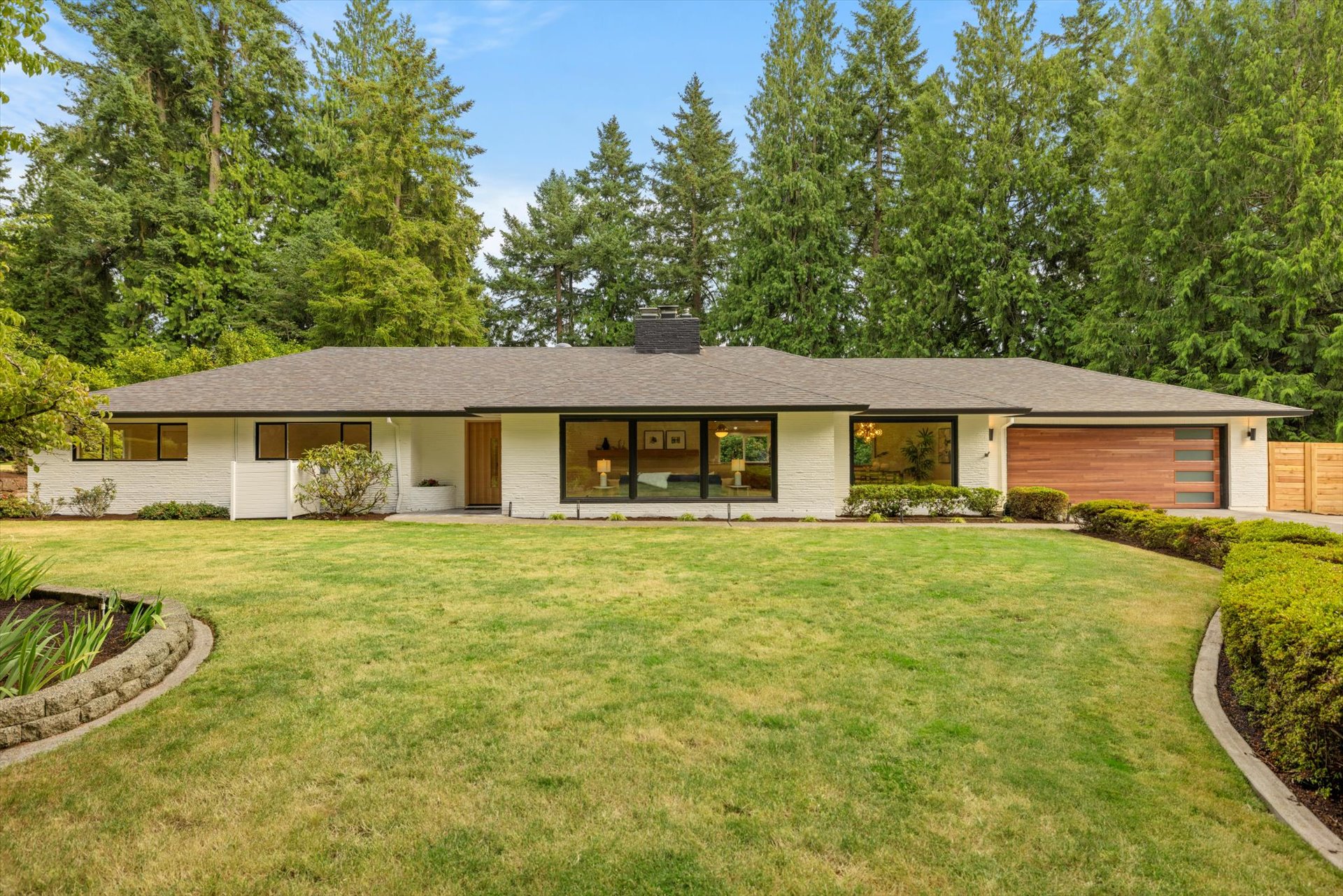 Single-story house with large front windows, surrounded by a green lawn and trees, under a blue sky.