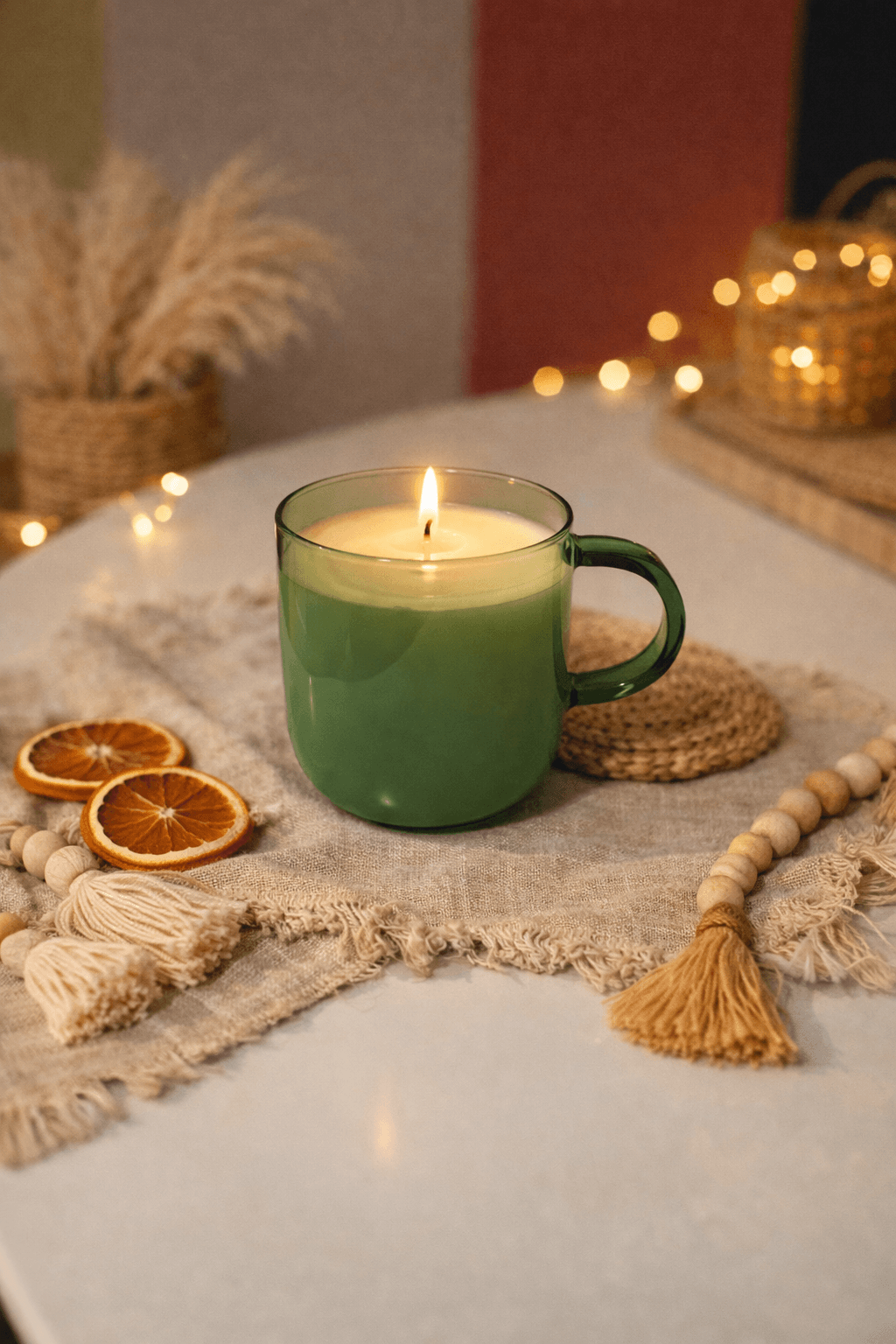 Green candle in a mug with dried orange slices, cookies, and decorative items on a textured cloth, warm background lighting.